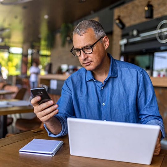 older man looking at his phone with his laptop open in front of him