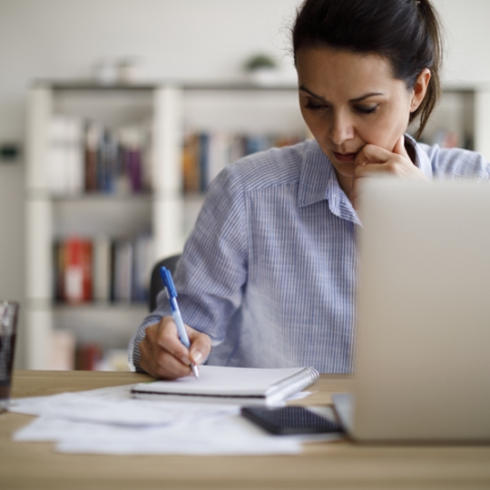 woman wearing button-up shirt writing in a notebook with a laptop in front of her