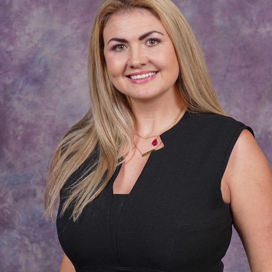 A blonde women in black dress smiling with a purple background behind her