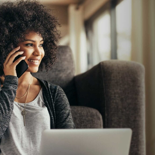 Women sitting down in from of laptop behind her is a couch. She is holding a phone smiling and laughing